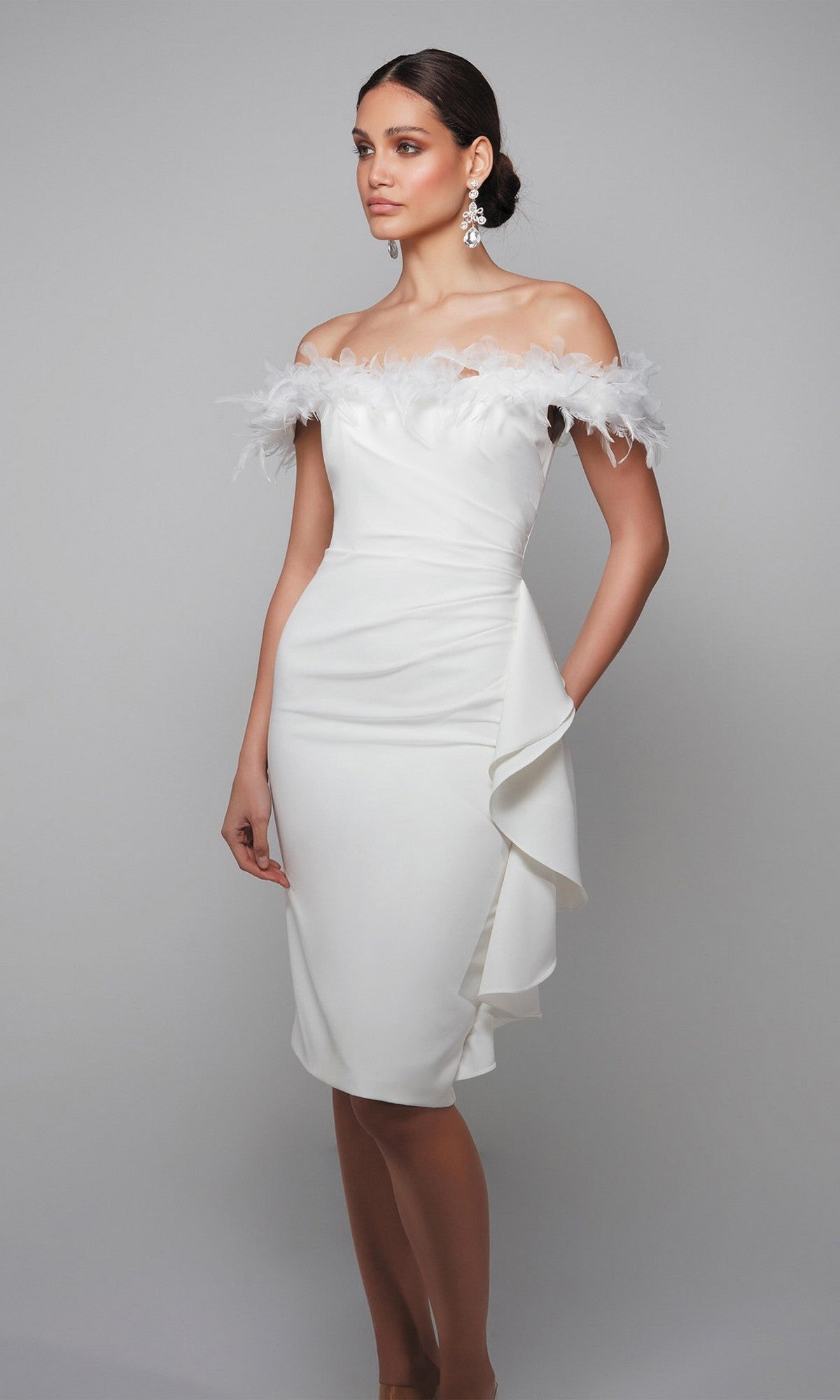 A woman stands against a plain gray background wearing the Alyce 70000 knee-length semi-formal dress, an off-the-shoulder white design featuring feather trim and a side ruffle.